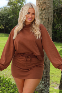 A woman wearing a brown matching two piece set featuring a cropped sweatshirt and mini skirt of the same material, standing outdoors with trees and other greenery in the background. 