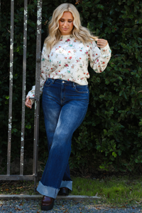 Woman model wearing an off-white sweater that has a multi-colored floral print pattern throughout with dark denim jeans. The photo is taken next to an iron gate outdoors with greenery in the back. 