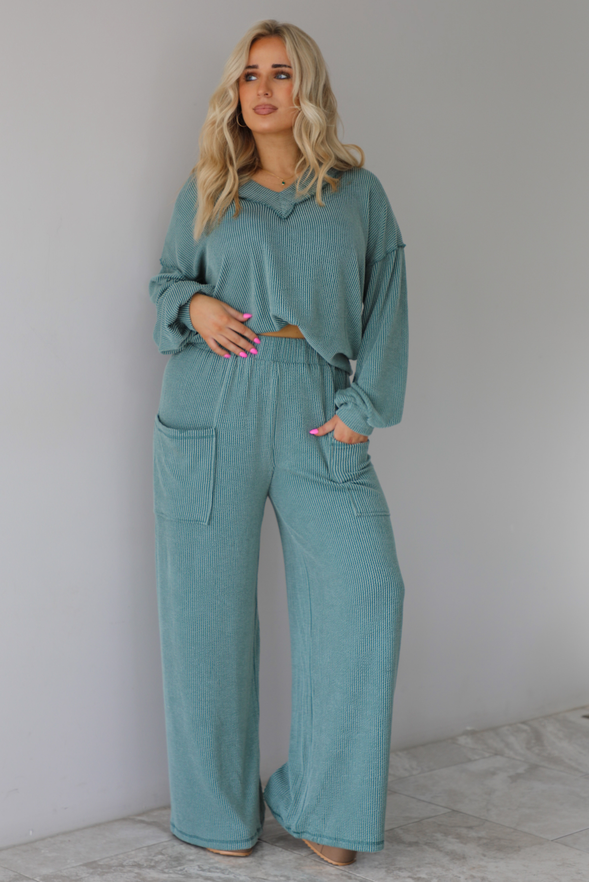 Woman wearing a teal two piece set that has a v-neck long sleeved oversized top and high waisted pants. The top is tucked into the woman's bra to give it a cropped look. The photo is taken indoors against a white wall with white marble flooring. 