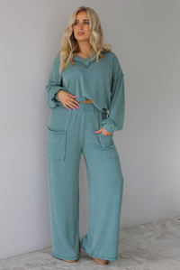 Woman wearing a teal two piece set that has a v-neck long sleeved oversized top and high waisted pants. The top is tucked into the woman's bra to give it a cropped look. The photo is taken indoors against a white wall with white marble flooring. 