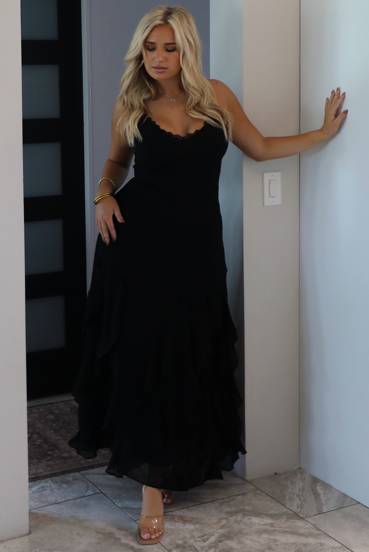 A woman wearing a black formal maxi dress with lace trim detailing at the bodice and ruffled details at the skirt, standing indoors in an archway with white marble flooring and a black door in the background. 