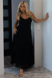 A woman wearing a black formal maxi dress with lace trim detailing at the bodice and ruffled details at the skirt, standing indoors in an archway with white marble flooring and a black door in the background. 