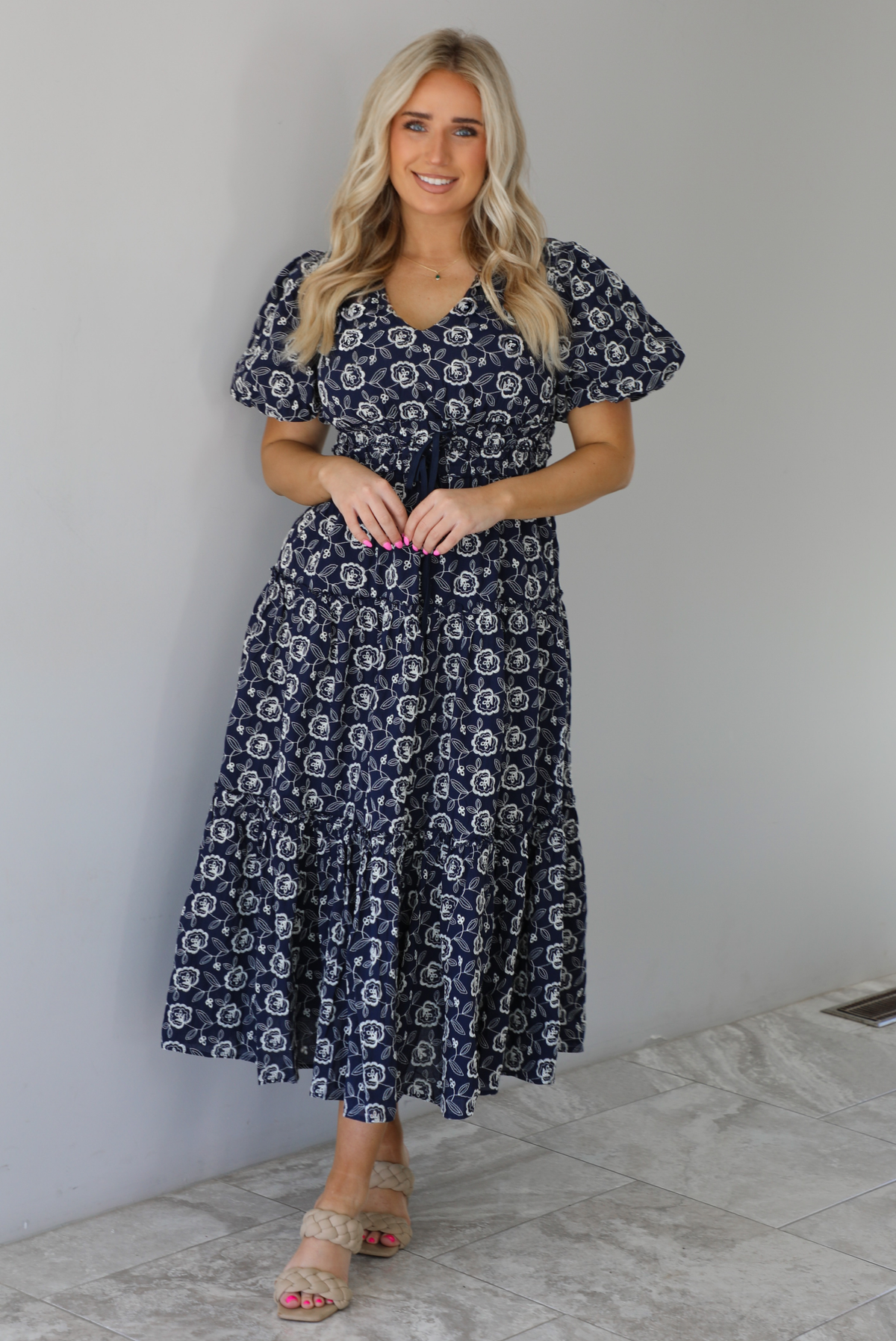 Woman wearing a navy dress that has a white embroidered floral print pattern throughout, a cinched waistline, and flowy maxi length skirt. The photo is taken indoors against a white wall with white marble flooring. 