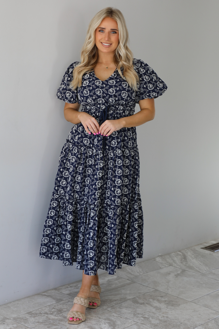 Woman wearing a navy dress that has a white embroidered floral print pattern throughout, a cinched waistline, and flowy maxi length skirt. The photo is taken indoors against a white wall with white marble flooring. 