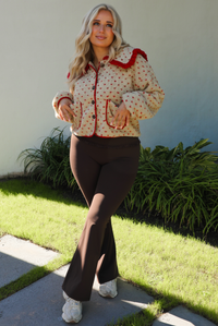 Woman model wearing a tan and red sherpa cardigan with a collared neckline and red trim with dark brown ahtleisure pants. The photo is taken outdoors with a white wall and greenery in the back. 