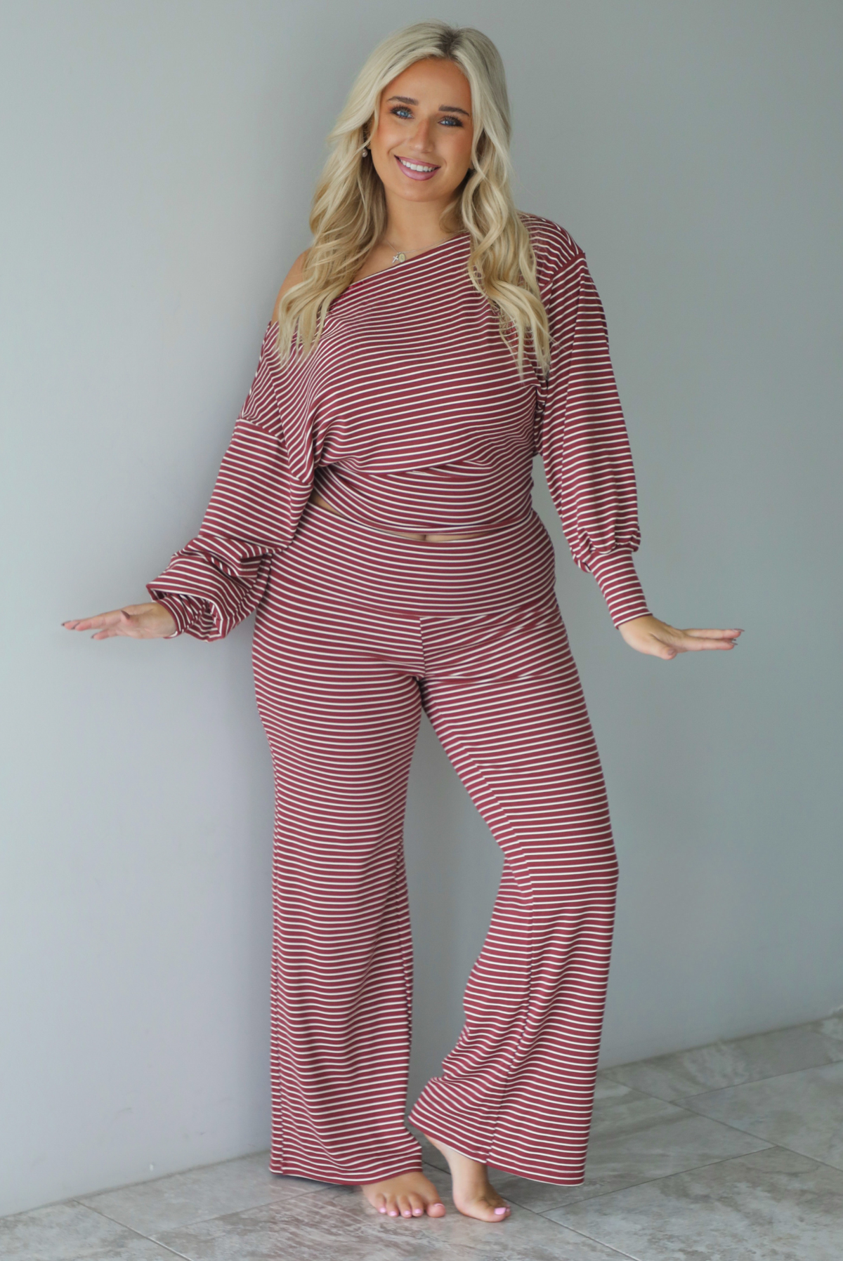 A woman wearing a maroon and white striped two piece matching set featuring a slouchy off the shoulder long sleeved top and mid-rise, folded over waistline wide leg pants, standing indoors against a white wall with white marble flooring. 