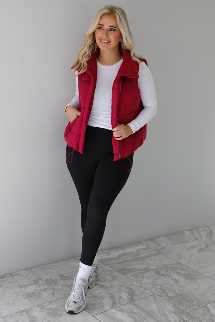 Woman wearing a wine red colored puffer vest, white long-sleeve shirt, black leggings, and gray sneakers with white ankle socks indoors against a white wall with white marble flooring.