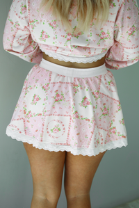 Woman wearing a two piece set featuring a pink floral set that has an oversized long sleeved top and high waisted skirt. The photo shows a closeup of the back of the skirt. The photo is taken indoors against a white wall.  