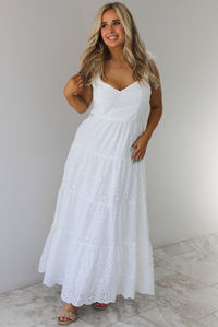 Woman wearing a white maxi length dress that has an eyelet floral design throughout and has a fitted bodice. The photo is taken indoors against a white wall with white marble flooring. 