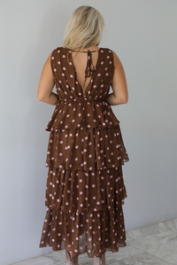 A woman shown from behind wearing a brown midi length ruffled dress with a pink polka dot print pattern throughout, standing indoors against a white wall with white marble flooring. The back of the dress shows a tie closure at the top of the dress. 