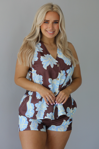Woman wearing a brown, blue, and off white floral two piece set featuring a vest style top with functional button closures in the front and shorts. The photo is taken in front of a white wall.