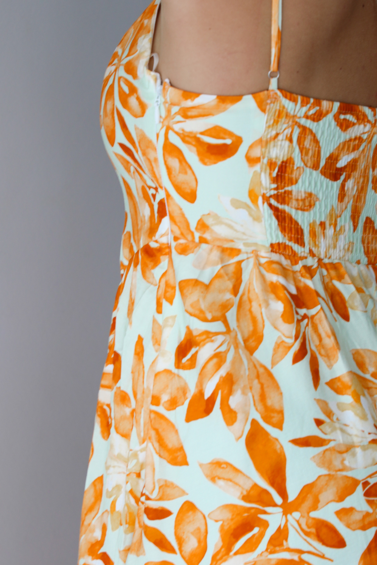 A woman shown from behind modeling a spaghetti strapped, floor-length maxi dress with a white base and a vibrant marigold orange floral pattern, standing indoors against a white wall. The photo shows the side of the dress, focusing on the zipper closure on the side. 