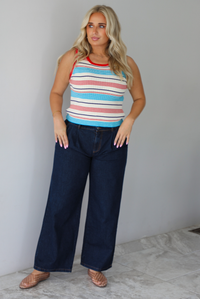 A woman modeling a sleeveless ribbed knit top with horizontal stripes in red, light blue, navy, and white, paired with dark wide-leg jeans and metallic rose gold flats, standing indoors against a white wall with white marble flooring. 
