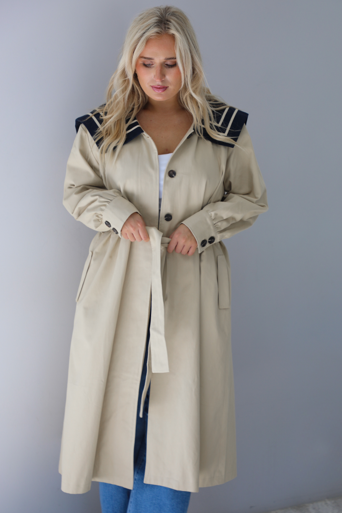 A woman wearing a tan trench coat with a navy collared neckline, functional button closures in the front, functional pockets on either side, with a tie waistline, paired with light wash flared hem denim jeans, standing indoors against a white wall.