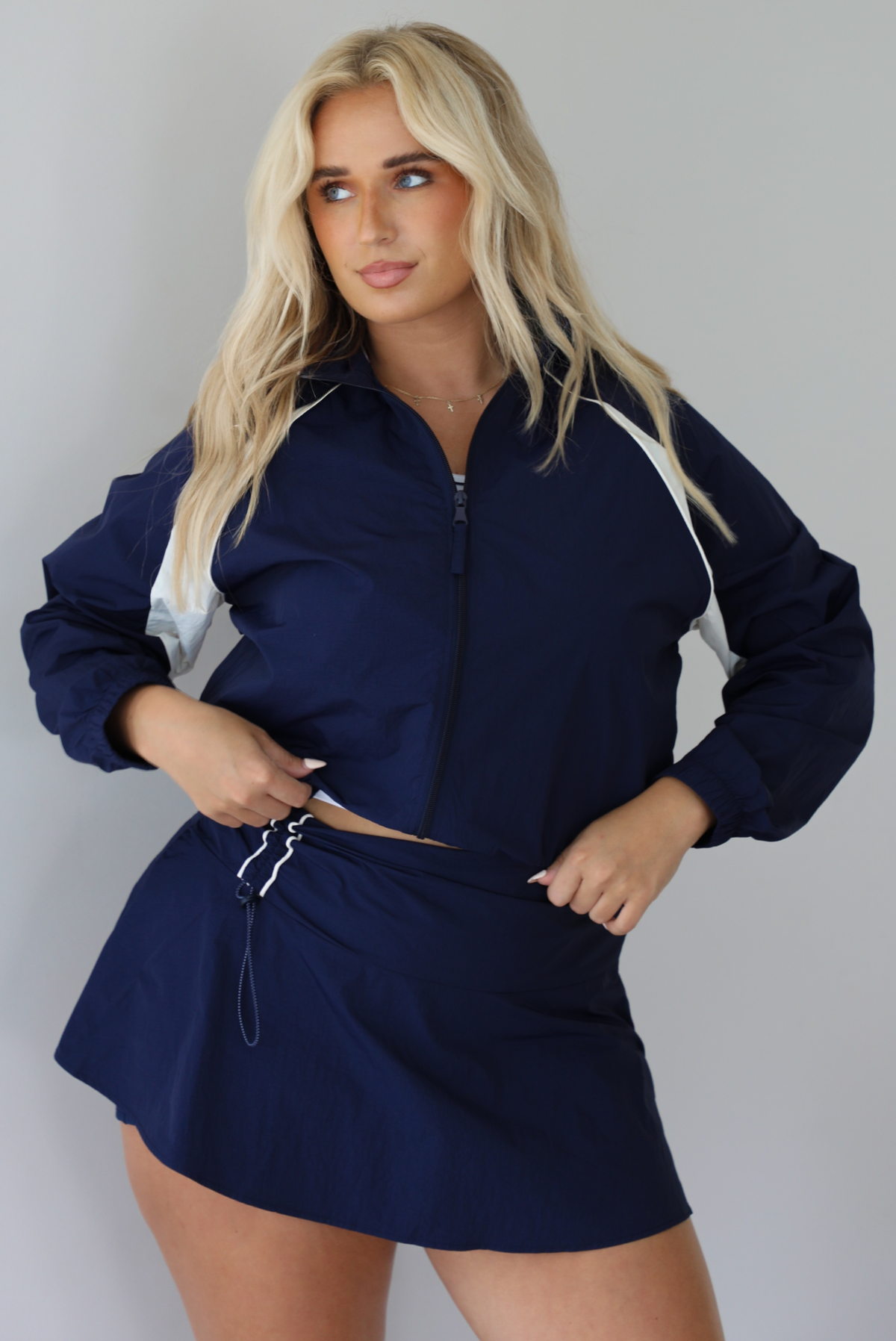 Woman wearing a navy tracksuit featuring a jacket and skort with a white tank indoors against a white wall. The photo shows the jacket zipped up.