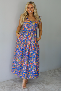 A woman wearing a sleeveless tiered navy blue maxi dress with a colorful floral print with a fitted bodice and flowy skirt with functional pockets on either side, standing indoors against a white wall with white marble flooring. 