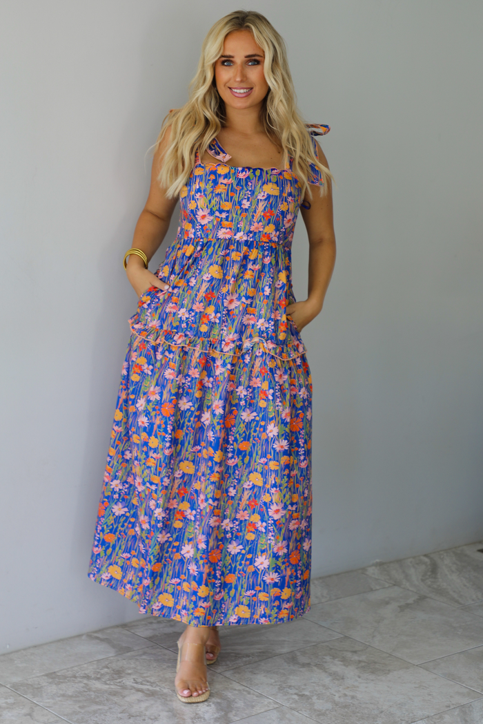 A woman wearing a sleeveless tiered navy blue maxi dress with a colorful floral print with a fitted bodice and flowy skirt with functional pockets on either side, standing indoors against a white wall with white marble flooring. 