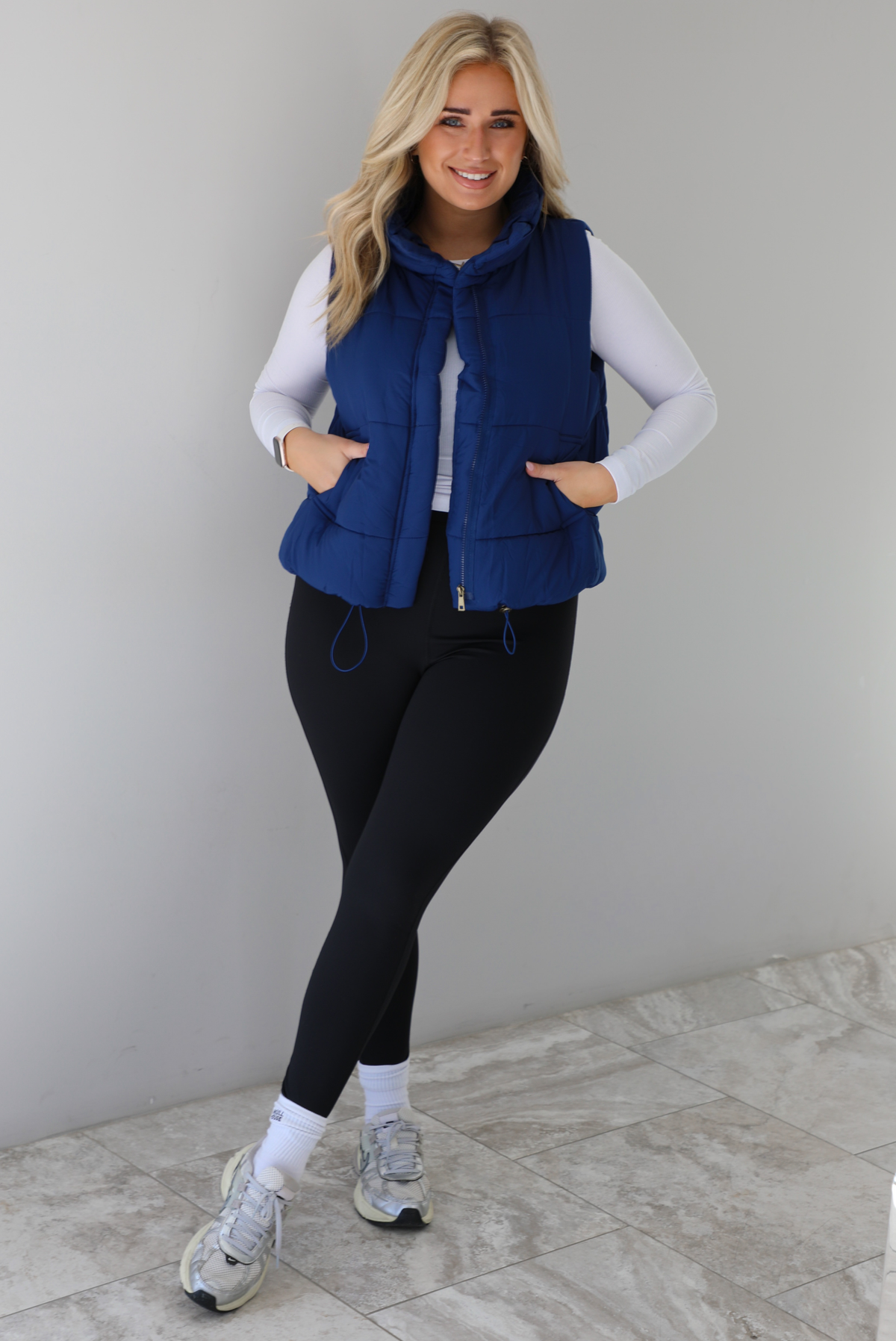 Woman wearing a navy colored puffer vest, white long-sleeve shirt, black leggings, and gray sneakers with white ankle socks indoors against a white wall with white marble flooring.