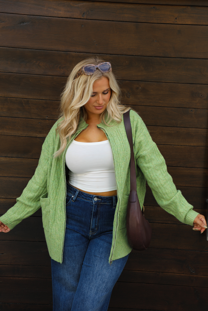 Woman model wearing a green cable knit zip-up cardigan with dark denim jeans outdoors against a dark brown wooden door. The cardigan is unzipped. The model is wearing a large.