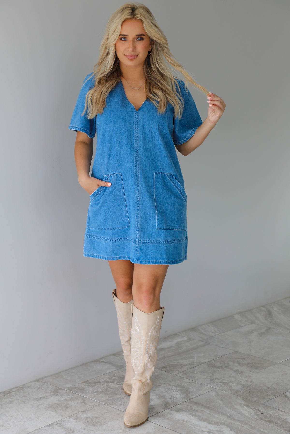 Woman wearing a v-neck, short sleeved, denim mini dress with functional pockets on either side. The photo is taken indoors against a white wall with white marble flooring. 