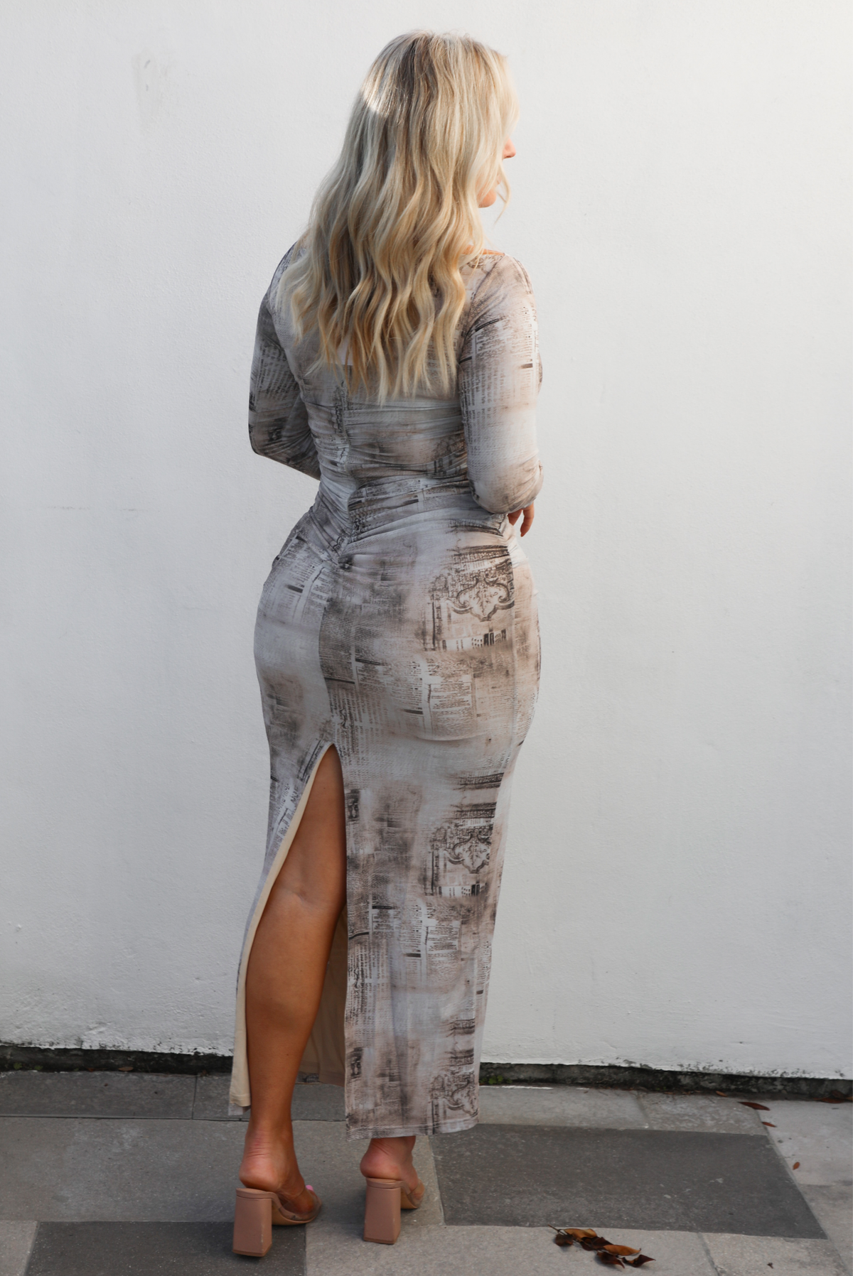 Back of a woman model wearing a multi-colored and multi-patterned maxi length dress that is fitted with long sleeves. The photo showcases the back of the dress, which has a zipper closure, and the split hem detail in the back. The photo is taken outdoors against a white wall. 