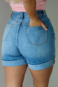 Woman wearing a medium wash pair of denim shorts that are high waisted, have functional front and back pockets, and a cuffed hem. The photo is taken indoors against a white wall and shows the back of the shorts. 