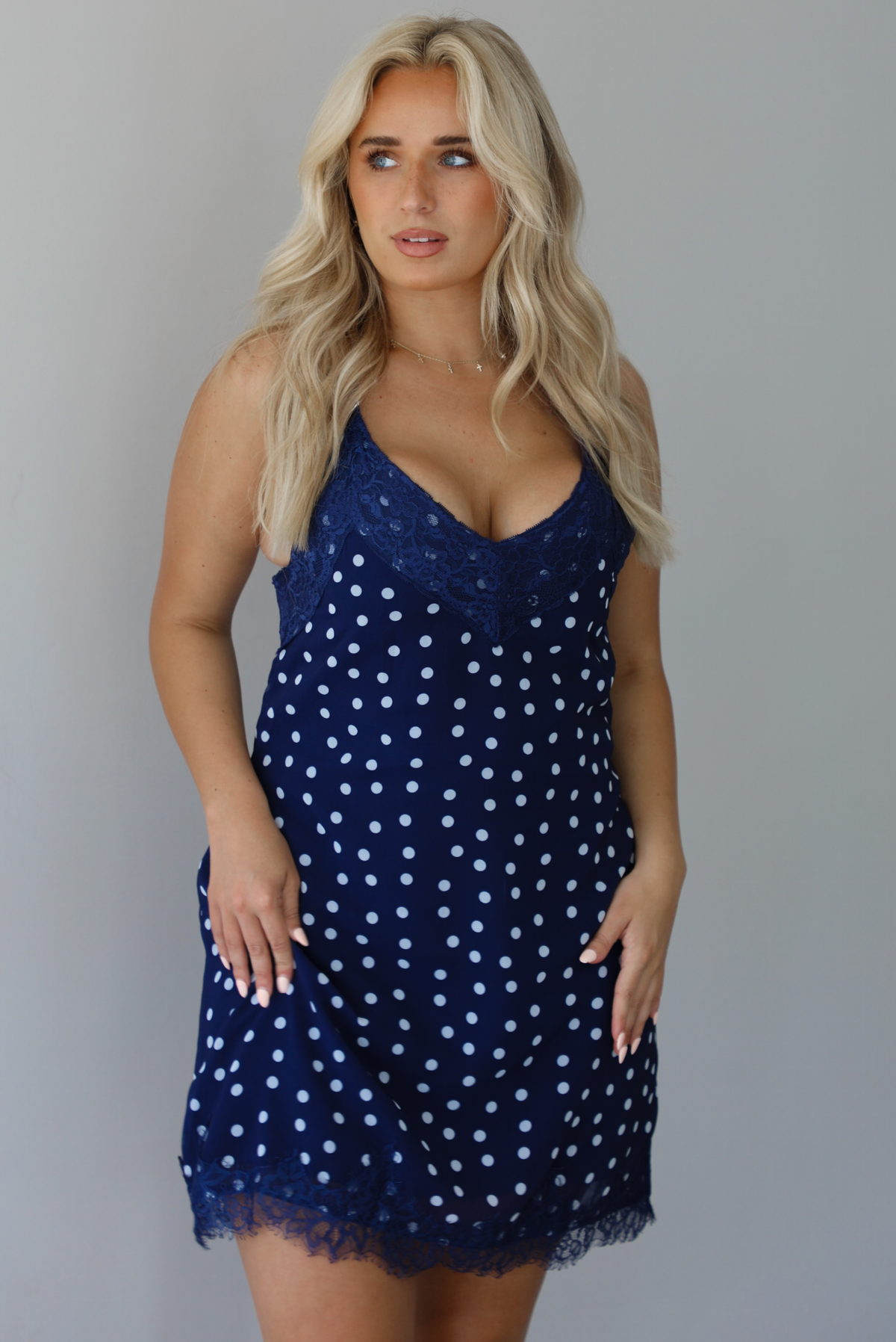 Woman wearing a navy dress with a white polka dot print pattern throughout with blue lace detailing at the neckline and hem indoors against a white wall.