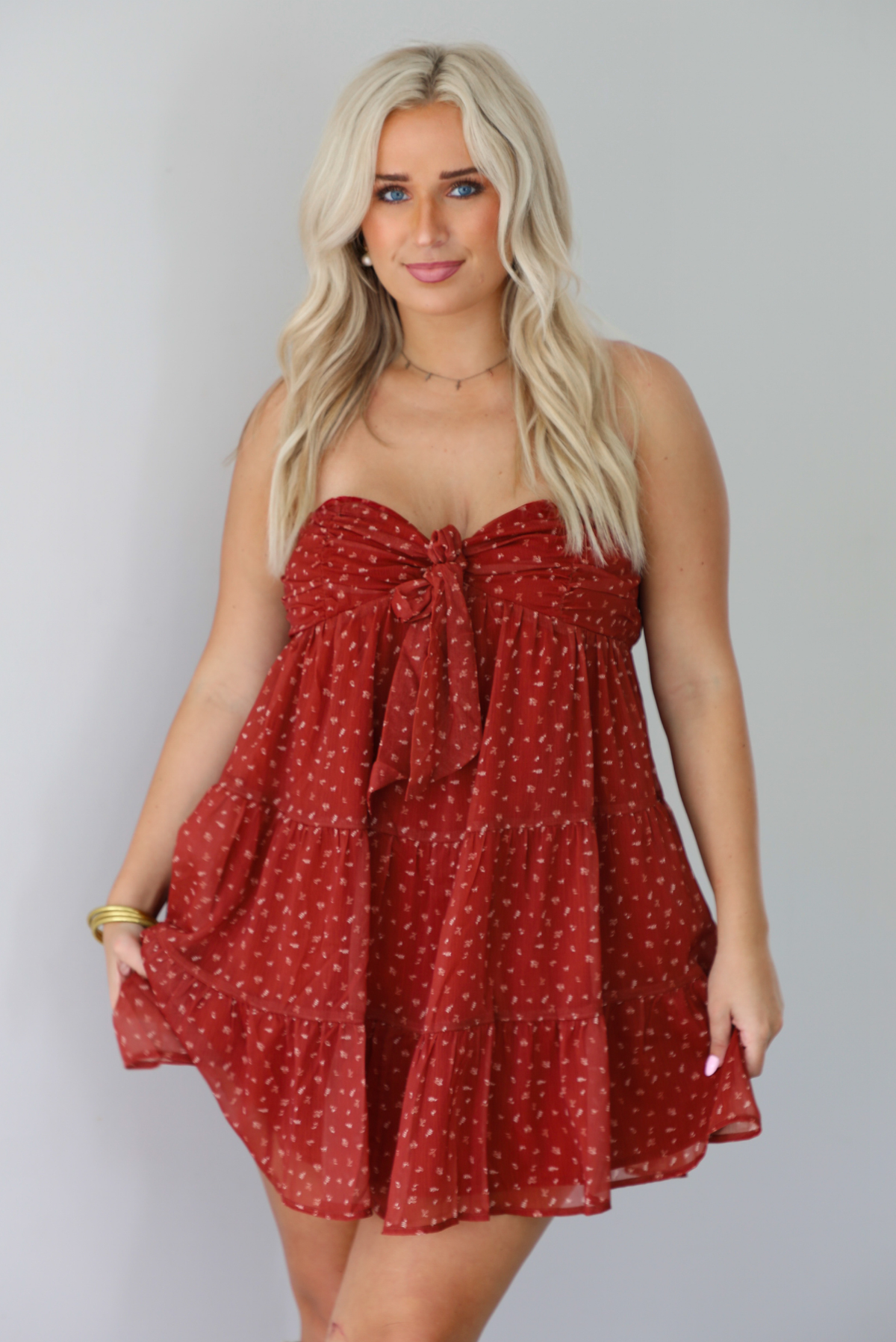A woman wearing a rust mini length dress with a cream floral and leaf tiny print design throughout, has a strapless design with a tie detail in the center of the bust, with a baby-doll dress cut that's fitted at the bust with a flowy skirt, standing indoors against a white wall.