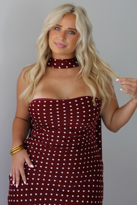 A woman wearing a strapless maroon midi dress with a white polka dot pattern with a matching scarf, standing indoors against a white wall. The photo shows a closeup of the bodice of the dress and the scarf. 