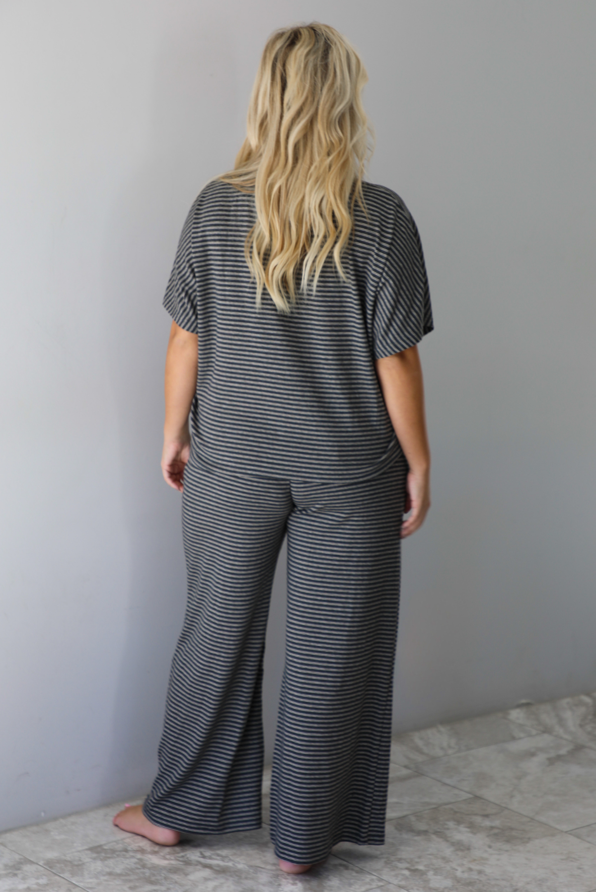 A woman shown from behind wearing a charcoal and white striped two piece matching set with green trim detailing at the neckline and waistline, featuring an oversized short sleeved top and high waisted pants, standing indoors against a white wall with white marble flooring. 