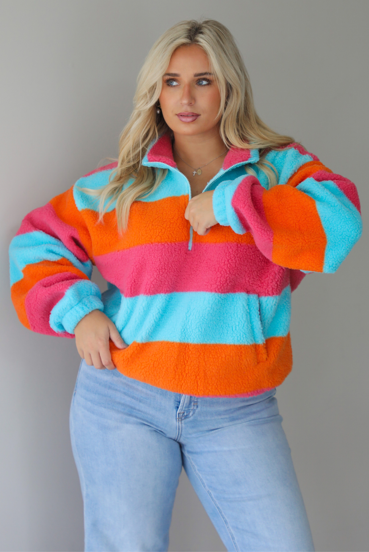 A woman wearing a turquoise, pink, and orange striped sherpa quarter-zip sweater with light wash denim jeans, standing indoors against a white wall.