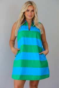 A woman wearing a sleeveless collared mini dress with bold horizontal stripes in green and blue, standing indoors against a white wall. The model is standing with their hands in the pockets on either side of the dress. 
