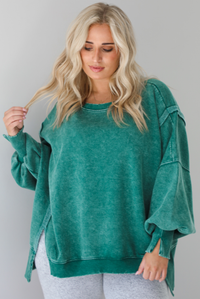 Woman wearing a green colored oversized sweatshirt with exposed seam details and a high-low design with heather grey casual pants. The photo is taken indoors against a white wall.