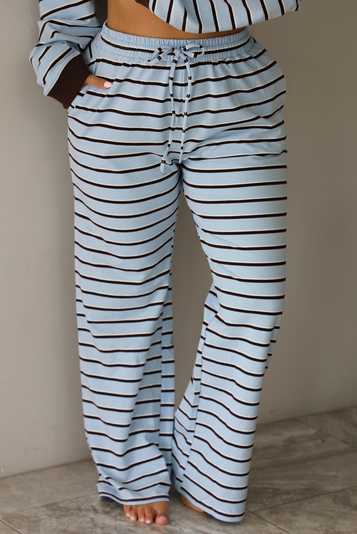 A woman wearing a blue matching two piece set with dark brown and white striped details, featuring an oversized long sleeved top and high waisted loose fitting, flared leg pants, indoors against a white wall with white marble flooring. The photo shows a closeup of the pants, which have an elastic and drawstring waistline and functional pockets on either side. 