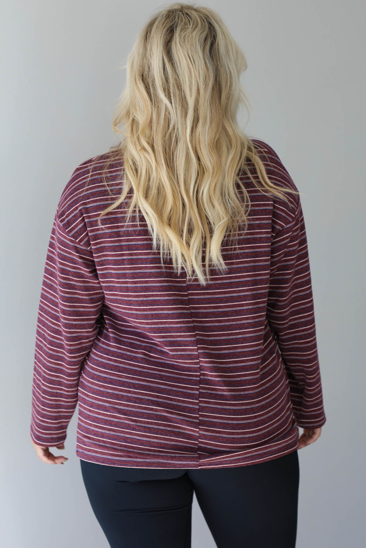 A woman shown from behind wearing a burgundy striped long sleeved top paired with black leggings, standing indoors against a white wall. 