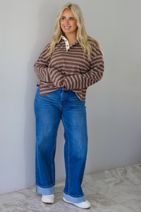 A woman wearing a brown and cream striped collared quarter-button long sleeved top, front tucked into dark wash denim wide leg jeans that have a cuffed hem, paired with cream sneakers, standing indoors against a white wall with white marble flooring. 