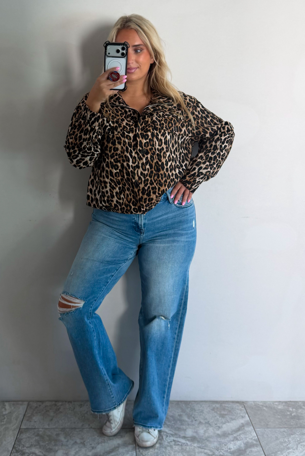 A woman wearing a leopard collared jacket paired with a pair of medium wash distressed denim jeans, standing indoors against a white wall with white marble flooring, taking a selfie. 