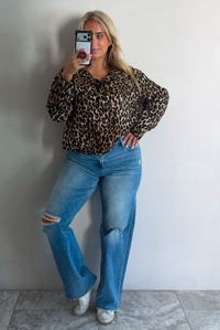 A woman wearing a leopard collared jacket paired with a pair of medium wash distressed denim jeans, standing indoors against a white wall with white marble flooring, taking a selfie. 