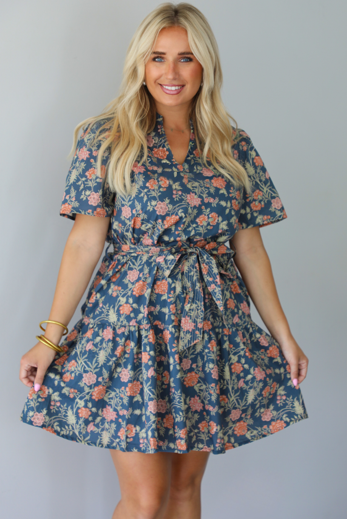 A woman modeling a short-sleeve floral mini dress with a tie waist, standing indoors against a white wall.