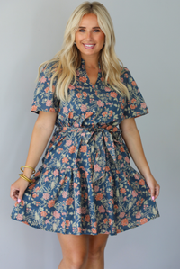 A woman modeling a short-sleeve floral mini dress with a tie waist, standing indoors against a white wall.