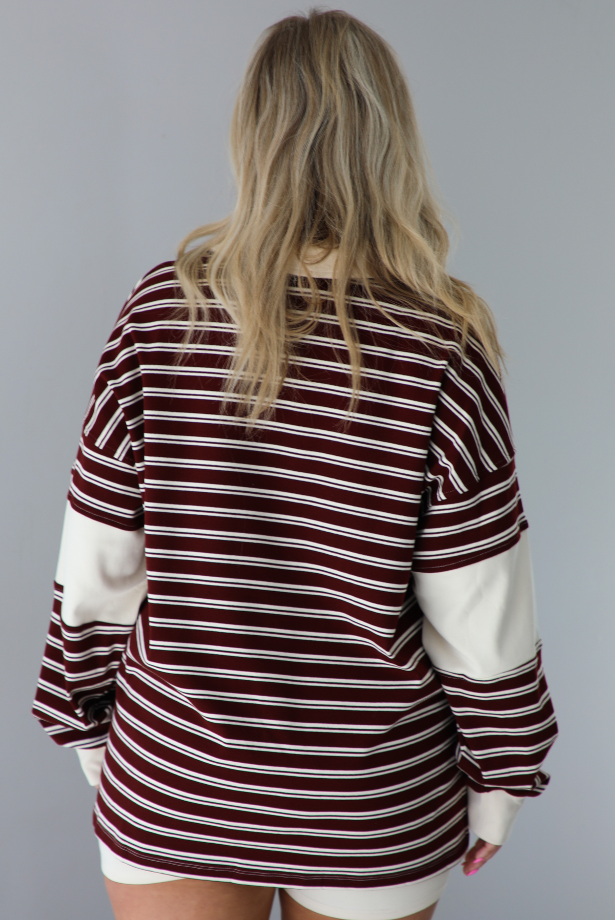 Woman wearing a burgundy and cream striped oversized collared top with cream colored biker shorts. The photo is taken indoors against a white wall. The photo shows the back of the top.