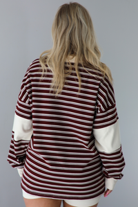 Woman wearing a burgundy and cream striped oversized collared top with cream colored biker shorts. The photo is taken indoors against a white wall. The photo shows the back of the top.