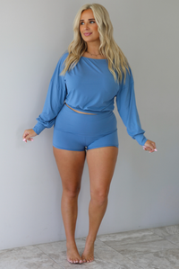 A woman wearing a dust blue matching two piece set consisting of a slouchy off the shoulder top that's fitted at the waist and folded over waist shorts, standing indoors against a white wall with white marble flooring. 
