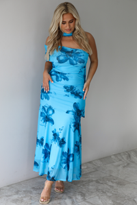 Woman wearing a blue floral maxi dress with a fitted bodice and flowy skirt indoors against a white wall with white marble flooring. 
