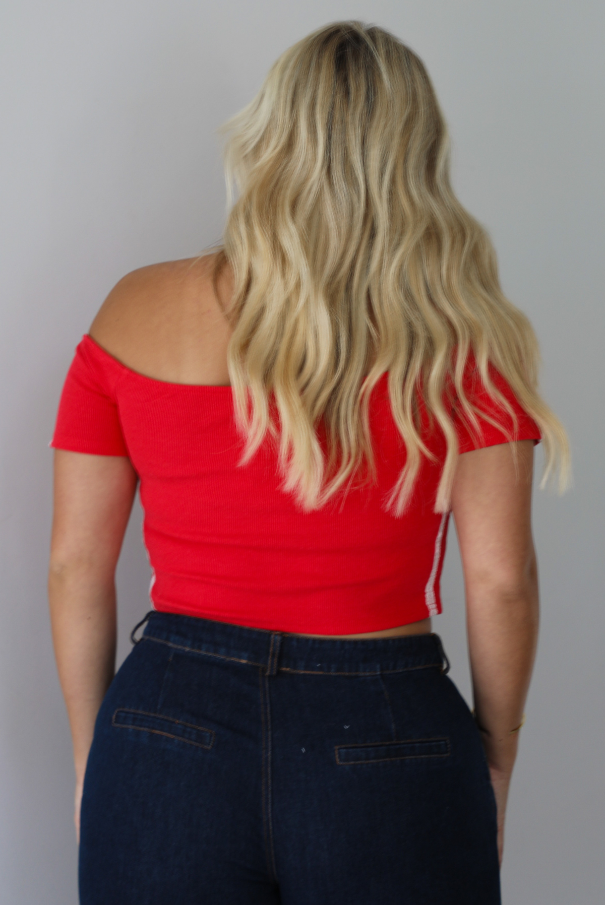 A blonde woman shown from behind modeling a bright red off-the-shoulder short-sleeve top with white stripes on the sleeves, paired with dark wide-leg denim jeans, standing indoors against a white wall.