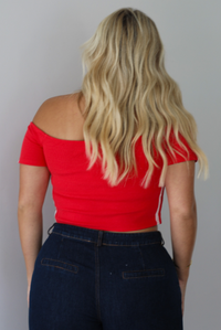A blonde woman shown from behind modeling a bright red off-the-shoulder short-sleeve top with white stripes on the sleeves, paired with dark wide-leg denim jeans, standing indoors against a white wall.