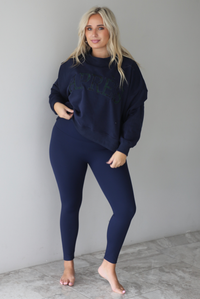 A woman wearing a navy sweater with Apreś written on the front in collegiate lettering in a plaid print, paired with a matching two piece navy set, standing indoors against a white wall with white marble flooring. 
