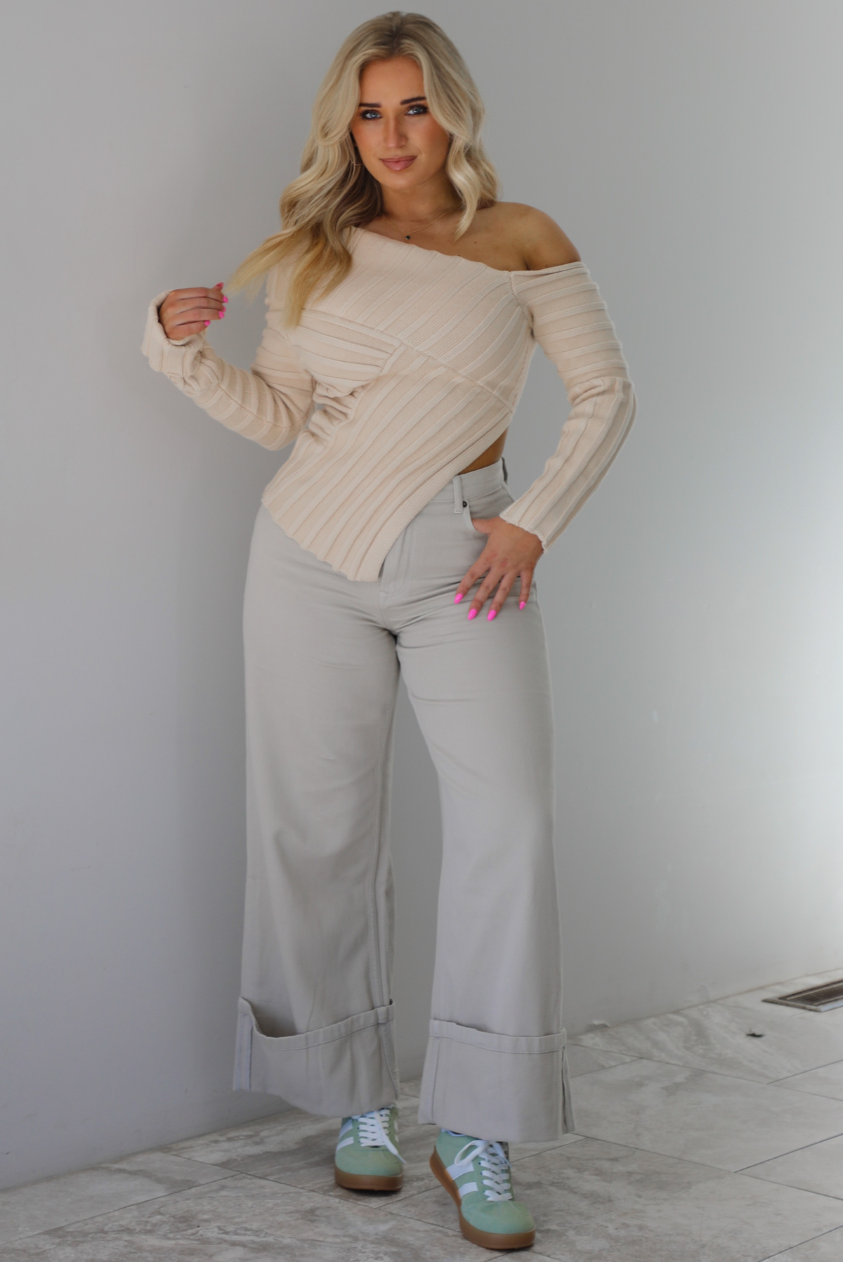 Woman wearing a cream colored ribbed textured knit top that has an asymmetrical hem with long sleeves in which one side is worn off the shoulder. The sweater is paired with sand colored high waisted, cuffed hem denim. The photo is taken indoors against a white wall with white marble flooring. 