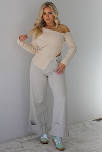 Woman wearing a cream colored ribbed textured knit top that has an asymmetrical hem with long sleeves in which one side is worn off the shoulder. The sweater is paired with sand colored high waisted, cuffed hem denim. The photo is taken indoors against a white wall with white marble flooring. 