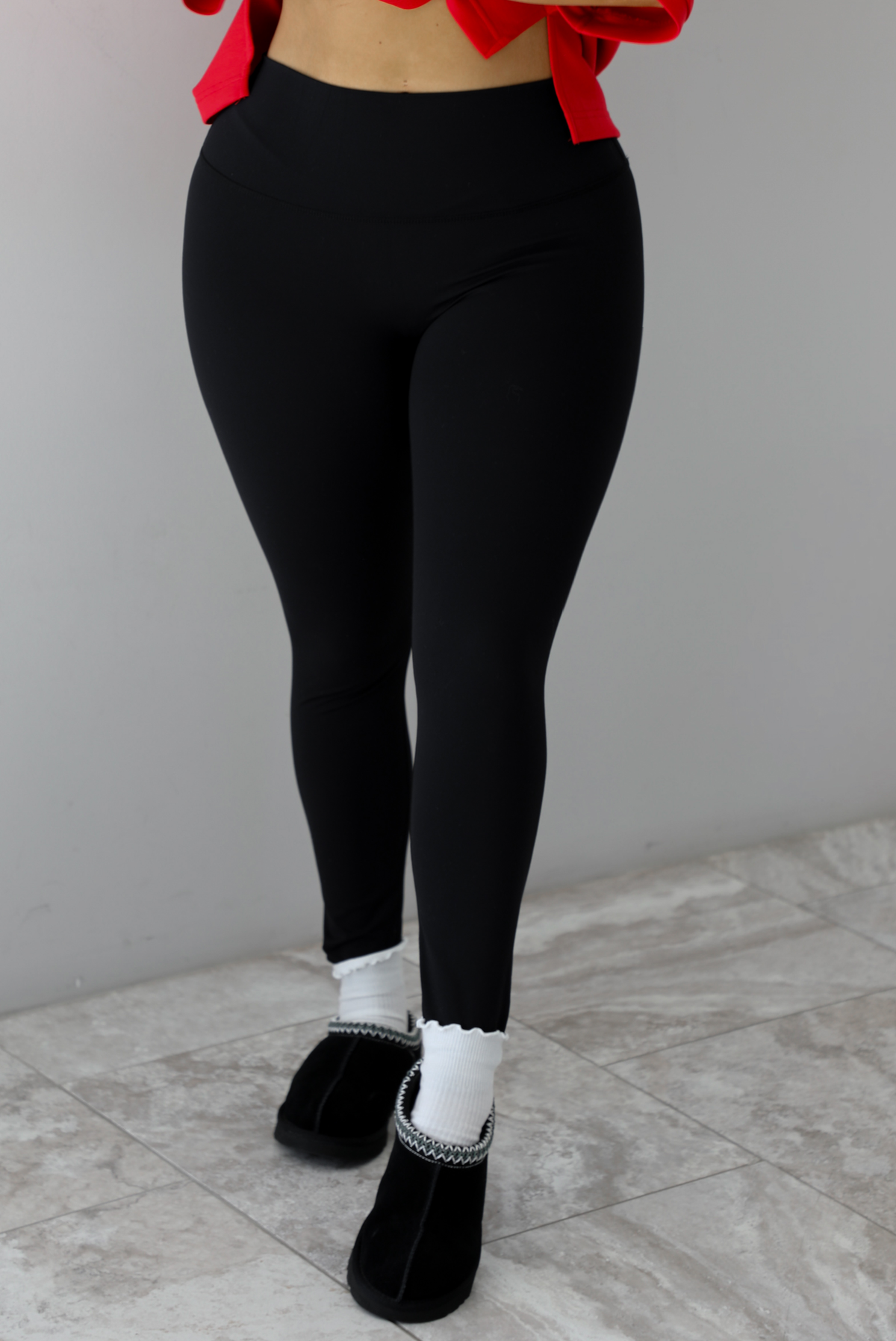Woman model wearing a fitted pair of black leggings that are high waisted, soft, and super stretchy. She's wearing them with a pair of white ruffled socks and black slip on ugg boot shoes. 